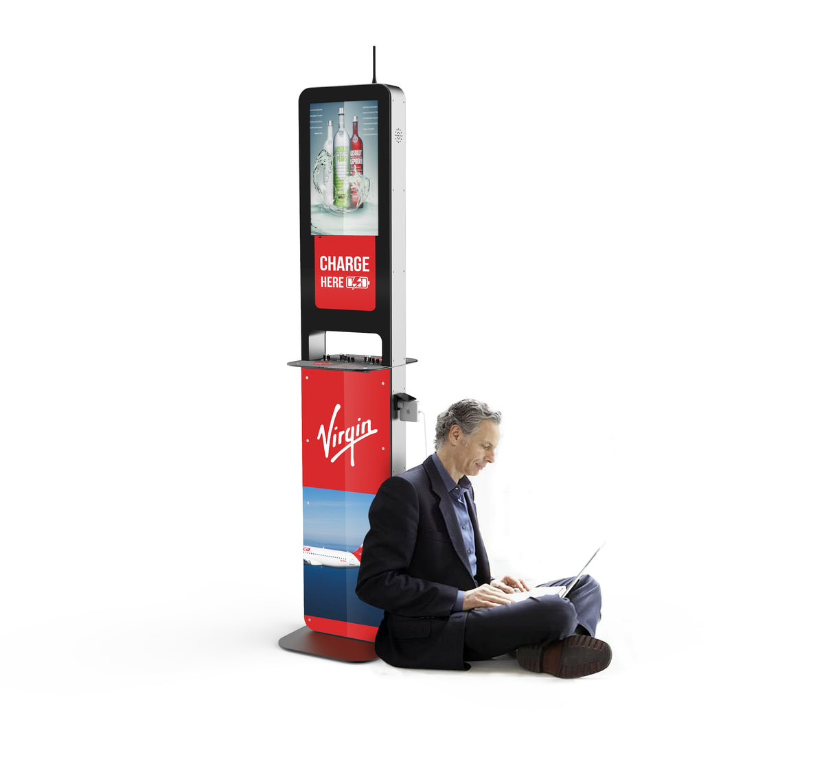 A man sits on the ground against the Aidan Charging Kiosk. His laptop's charger is plugged into the charging station behind him. Advertisements for liquor and Virgin Airlines are displayed on the screen of the kiosk. A sign stating "Charge Here" is displayed between both advertisements.