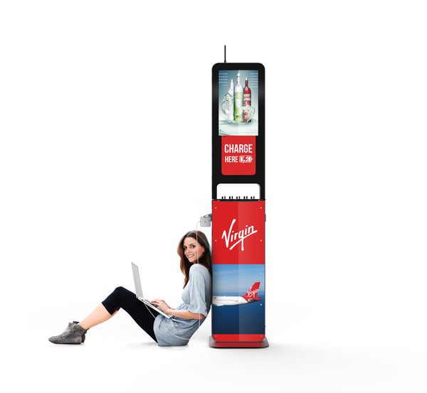 A woman sits on the ground against the Aidan Charging Kiosk and smiles as she uses her laptop.  The laptop's charger sits securely above the woman's head, plugged into both the Aidan charging kiosk and the computer on the woman's lap. A sign stating "Charge Here" is displayed between an advertisement for liquor and another for Virgin Airlines. 