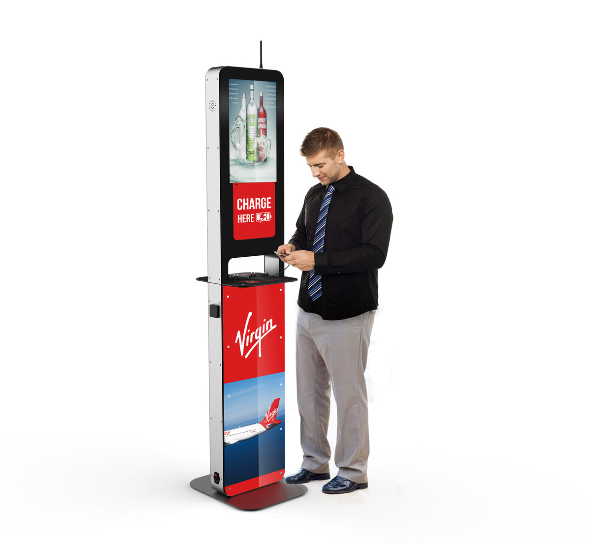 A man stands beside the Aidan Charging Kiosk with his cell phone plugged into the charging station. An advertisement for liquor sits above a sign that states "Charge Here" while another ad for Virgin Airlines sits at the bottom half of the phone charging station. 
